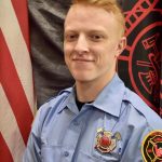 A firefighter with short red hair and a light smile poses in uniform in front of an American flag and a fire department flag. He wears a light blue shirt with patches and a badge. The background includes a dark cloth and the colorful flags.