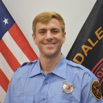 A smiling firefighter in a light blue uniform stands in front of the American flag and a black flag with text "DALE" partially visible. The firefighter has short blonde hair and an embroidered badge on his right sleeve.