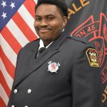 A firefighter in formal uniform, featuring a badge and insignia, stands in front of a backdrop with the American flag and a black flag with fire department insignia. He has a slight smile and wears a dark suit with silver buttons.