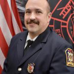 A firefighter with a mustache is posing for a portrait. He is wearing a navy blue uniform with fire department insignia on the badge and shoulder patch. An American flag and a fire department flag are in the background.