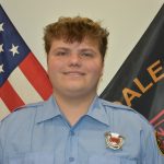 A firefighter in a light blue uniform shirt is standing in front of an American flag and a black flag. The individual has short, curly hair and is smiling at the camera. The shirt features a badge and patches on the sleeves.