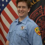 A person smiling while wearing a light blue uniform with badges, standing in front of an American flag and another flag with firefighting symbols.