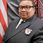 A person in a dark uniform wearing glasses and a badge, standing in front of an American flag and a black banner with red and gold detailing. The person has a short haircut and appears to be posing for a formal photograph.