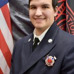 A young man wearing a navy uniform adorned with a silver badge and ribbon, standing in front of an American flag and what appears to be a dark colored flag with an emblem and red text. He is smiling at the camera.