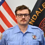 A firefighter wearing a light blue uniform and glasses stands in front of an American flag and a black flag with red details. The firefighter has a mustache and short brown hair. The flags partially cover a white wall in the background.