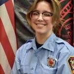 A person with shoulder-length hair and glasses is wearing a light blue uniform with an embroidered badge. They are smiling and standing in front of the American flag and a dark-colored flag featuring a red emblem.