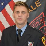 A young firefighter, dressed in a formal uniform with a badge, stands in front of an American flag and a black flag featuring fire department insignia and text. He has short, light hair and a serious expression.