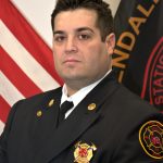 A firefighter in formal uniform poses for a photo. He stands in front of a backdrop featuring an American flag and a flag with the text "Glendale" partially visible. The uniform bears insignias, including a badge with the fire department's emblem.