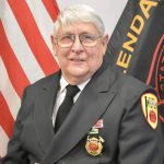 An older gentleman in a dark formal fire service uniform with badges and insignia stands in front of American and fire department flags. The man has white hair, glasses, and a friendly expression.
