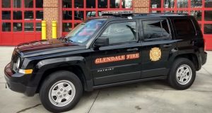 A black Jeep Patriot with "Glendale Fire" and a fire department logo on the side is parked in front of a fire station with red garage doors. The vehicle has emergency lights mounted on the roof.