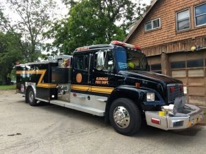 A black and yellow fire truck labeled "Glendale Fire Dept" is parked outside a two-story building with brown shingles and a garage door. Trees and greenery are visible in the background. The fire truck has various equipment compartments.