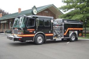 A Glendale Fire Department fire engine is parked in front of a brick building with a clock mounted on the roof. The vehicle features various gauges and hoses, with "45" marked on its side. A tree is visible on the right side of the image.