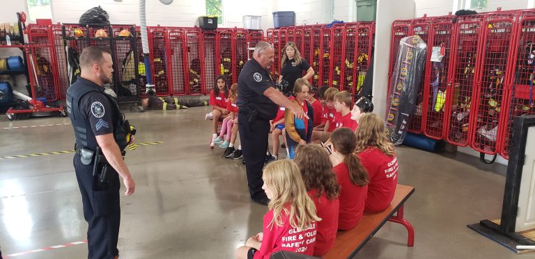 A group of children wearing red shirts are sitting on benches in a fire station, engaged in conversation with two uniformed police officers. The background features open lockers with firefighting gear. The scene appears to be an educational or community event.