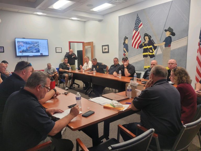 A group of around 14 people, predominantly men, sit around a conference table in a meeting room. A large mural depicting firefighters raising an American flag is on the wall. Papers and water bottles are scattered on the table, and a monitor shows a map on the left.