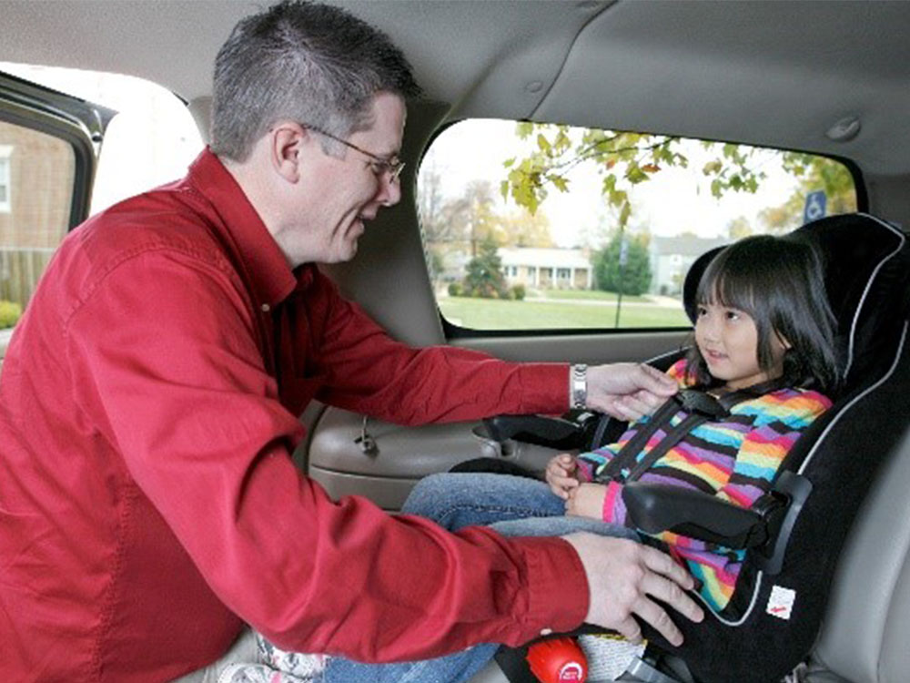 An adult wearing a red shirt is securing a smiling child in a colorful striped sweater into a car seat. They are inside a vehicle, with a view of trees and a building visible through the window.