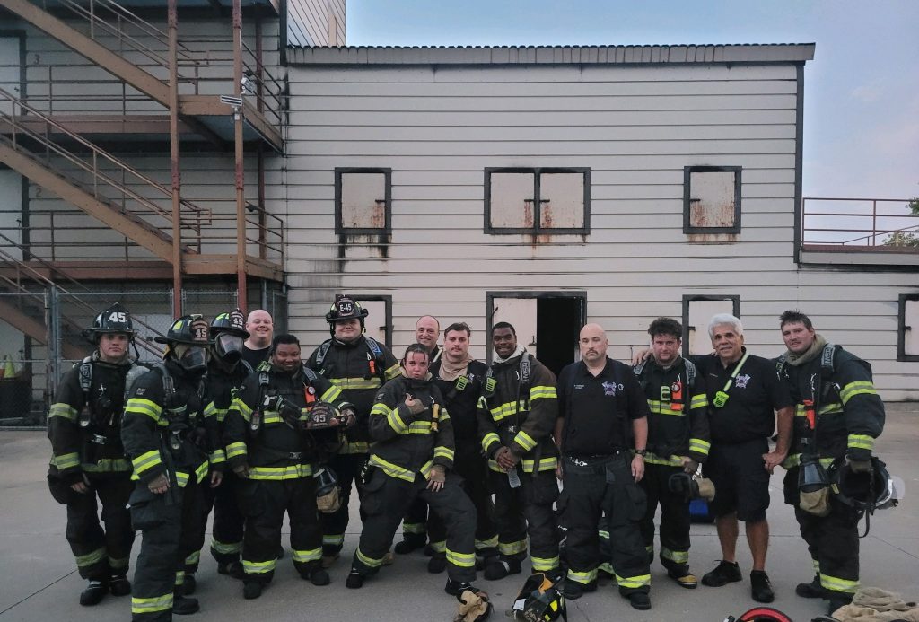 A group of firefighters in uniform pose for a photo in front of a training building. Some are in full gear with helmets, while others wear black shirts and pants with reflective strips. The building behind them is two stories tall with boarded-up windows.